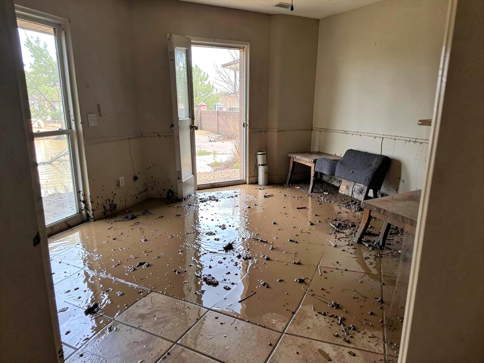 Before flood cleanup — living room with several inches of muddy brown floodwater across the tile, silt and debris floating, mud line on the walls, photographed from the doorway.