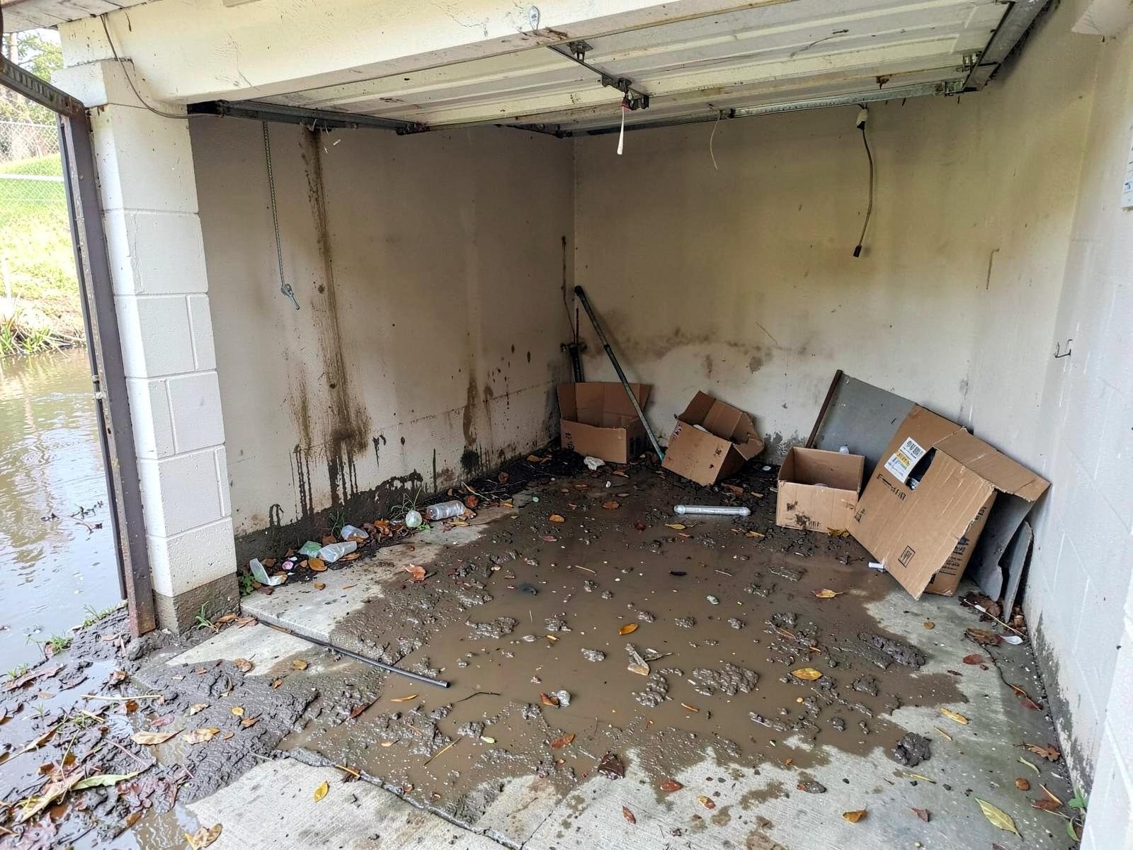 Before flood cleanup — residential garage with muddy stormwater and silt across the concrete slab, soaked cardboard boxes, leaves and debris washed in under the door, waterline stain on the wall.