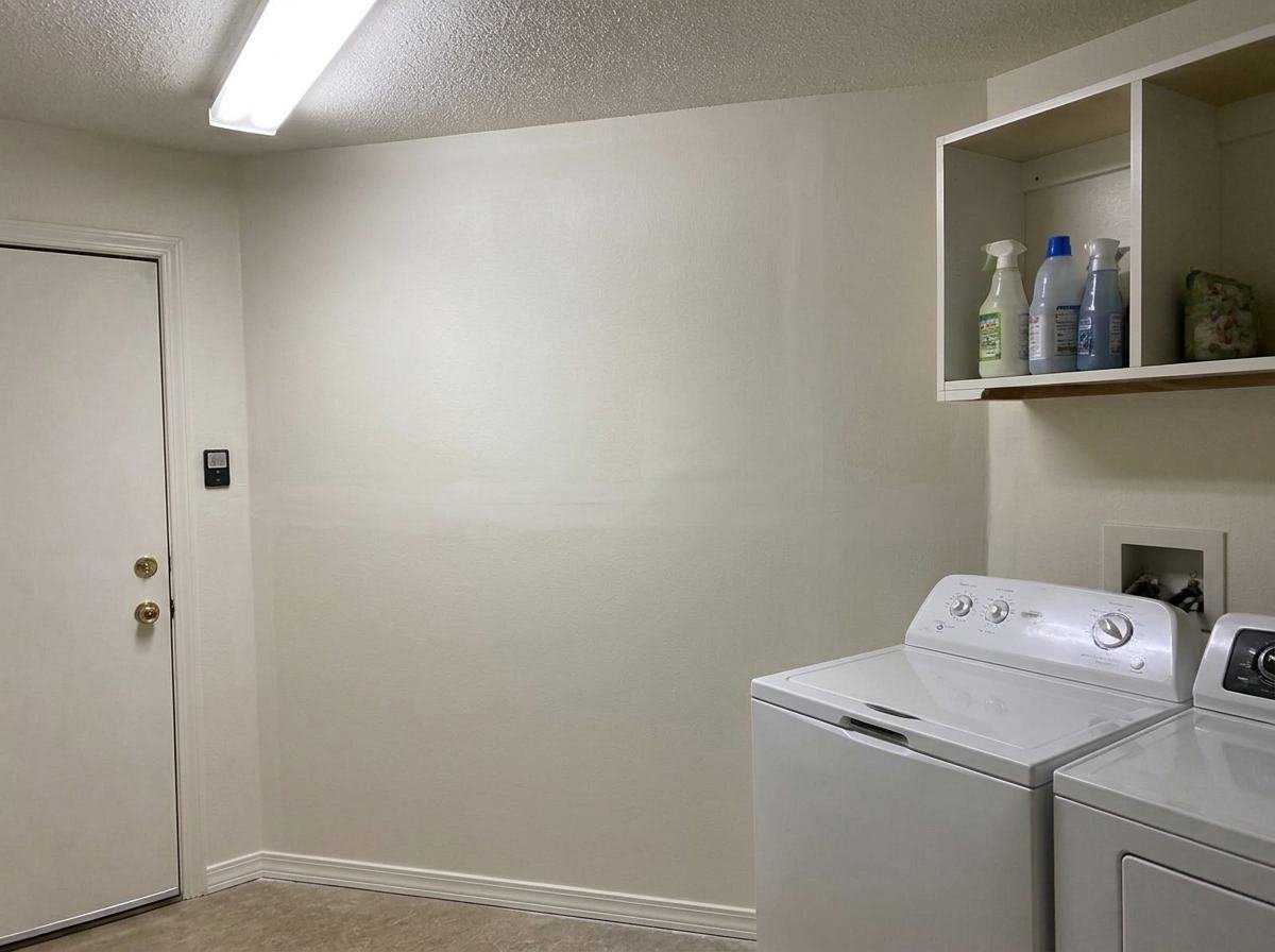 Job #WD-2503-029 after restoration — same laundry room, same camera angle as the before photo. Wall fully rebuilt, freshly painted clean off-white, new baseboard, ceiling clear, washer and dryer back in place.
